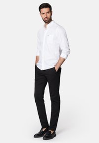 Biała koszula z guzikami i kieszenią na piersi, w połączeniu z czarnymi spodniami. Model ma na sobie czarne mokasyny. Materiał wydaje się gładki i wyrazisty.