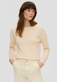 Pull en tricot beige clair à manches longues, décolleté large, et coupe droite. Porté avec un pantalon beige clair à fermeture à double bouton.