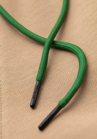 Zelené zapletené tkaničky s černými plastovými konci leží na béžovém textilním povrchu. Tkaničky mají hladkou strukturu a odolný design.