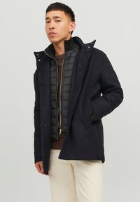 Abrigo exterior negro con cuello alto, botones y bolsillos con solapa, superpuesto a una chaqueta interior negra acolchada. Se lleva con pantalones de color claro.