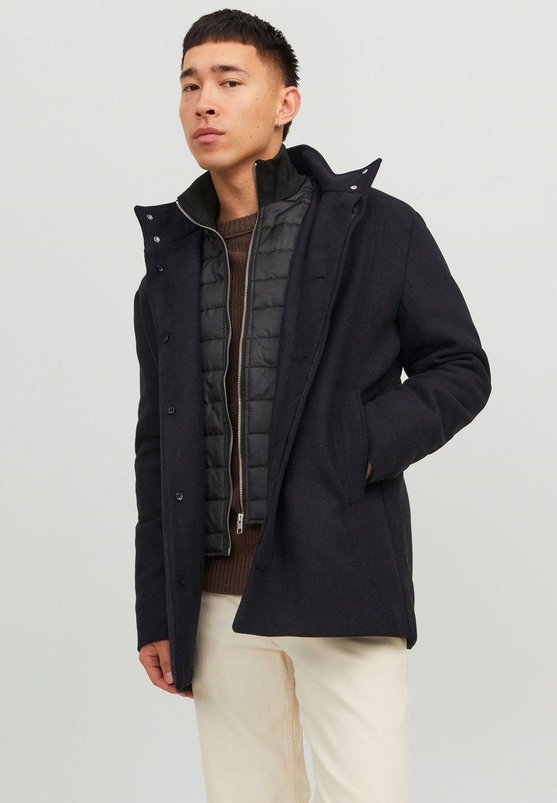 Abrigo exterior negro con cuello alto, botones y bolsillos con solapa, superpuesto a una chaqueta interior negra acolchada. Se lleva con pantalones de color claro.