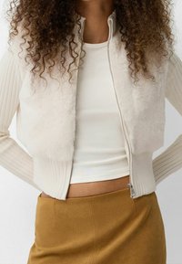 Bílá vesta na zip z umělé kožešiny s žebrovanými texturovanými rukávy. Nosit se může přes bílý top, v kombinaci s hnědou sukní, ukazující vrstvený styl.