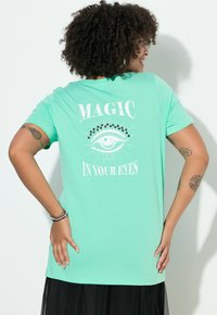 Mátově zelené tričko s krátkým rukávem s grafickým motivem a nápisem "MAGIE VE TVÝCH OČÍCH" a ilustrací oka na zádech.