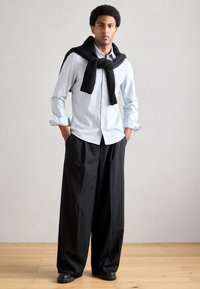 Ανοιχτό μπλε πουκάμισο με κουμπιά, μαύρο πλεκτό πουλόβερ δεμένο γύρω από τον λαιμό, μαύρο παντελόνι με φαρδιά πόδια και μαύρα παπούτσια σε ξύλινο πάτωμα.