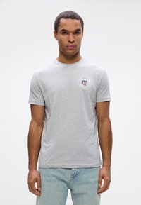 Grå bomulds t-shirt med rund halsudskæring og korte ærmer. Bemærkelsesværdig rundt logo på venstre bryst med røde og blå accenter.