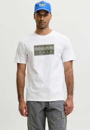 Mand iført hvid T-shirt med Jack & Jones-logo, grå cargobukser og en blå kasket, står mod en ensartet lys baggrund.