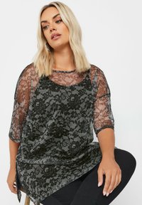 Top in pizzo nero con maniche trasparenti, pattern floreale, scollatura rotonda e spacchi laterali. Indossato sopra un top nero, abbinato a pantaloni neri.