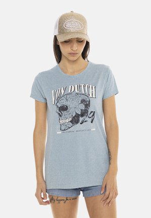 Femme portant un t-shirt Von Dutch bleu clair avec un design de crâne, un short en jean et une casquette Von Dutch dorée pailletée, debout devant un fond blanc.