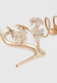 Kullast kõrge kontsaga sandaalid, kaunistatud lillemotiividega mis on tehtud kristallidest, õhukese rihmastiiliga ja läikiva, siledapinnalise viimistlusega.