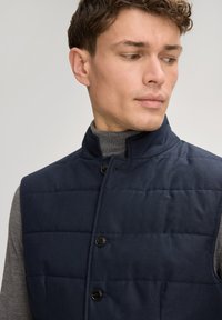 Granatowa pikowana kamizelka z wysokim kołnierzem i zapięciem na guziki z przodu; wykonana z gładkiej tkaniny o subtelnym wzorze, noszona na szarym swetrze.