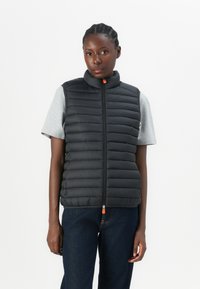 Black padded vest with high collar, zip closure featuring an orange pull tab, worn over a gray short-sleeve shirt, paired with dark jeans.