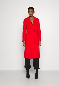Roter Wollmischmantel mit einem Gürtel, Reverskragen und langen Ärmeln; verfügt über Seitentaschen und eine gerade Silhouette. Getragen über schwarzen Hosen.