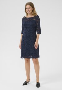 Abito in pizzo blu navy con maniche a tre quarti e orlo scallop. Presenta motivi floreali e scollatura a barca. Indossato con décolleté nere con fascia posteriore.