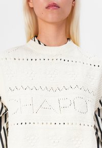 Knitted white sweater with openwork detailing and the text "CHAPO." Features a rounded neckline and layered with a black-and-white striped shirt underneath.