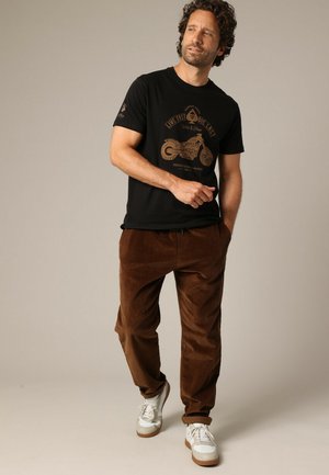 Zwarte t-shirt met gouden motorfiets graphic, bruine corduroy broek en witte sneakers met bruine zolen, staande voor een neutrale achtergrond.