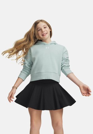 Ragazza sorridente con i capelli lunghi indossa una felpa azzurra con cappuccio e una gonna nera, in piedi con le braccia leggermente distese su uno sfondo semplice.