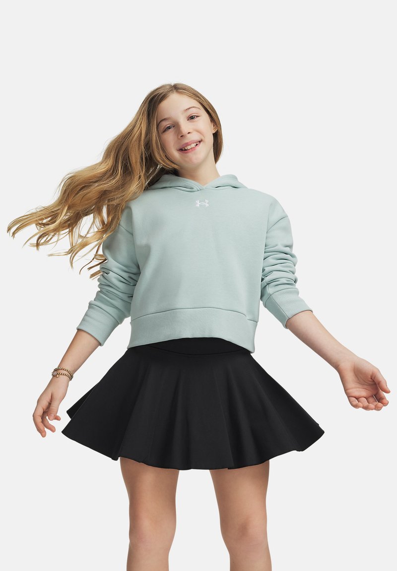 Fille souriante aux longs cheveux portant un sweat à capuche bleu clair et une jupe noire, debout les bras légèrement étendus devant un fond uni.