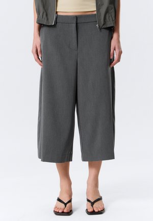 Donna che indossa pantaloni grigi larghi alla caviglia, sandali neri con punta quadrata, top beige corto e giacca verde scuro con zip, in piedi contro uno sfondo bianco.