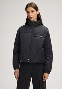 Schwarze gesteppte Jacke mit hohem Kragen, Reißverschluss und einer kleinen Reißverschlusstasche auf der Brust. Verfügt über elastische Bündchen und ein kurzes Design.