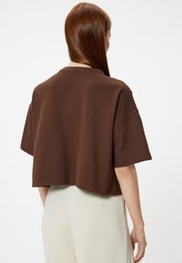 Brązowy krótki t-shirt z krótkim rękawem, wykonany z miękkiego materiału. Posiada luźny krój oraz widoczne szwy wzdłuż dołu.