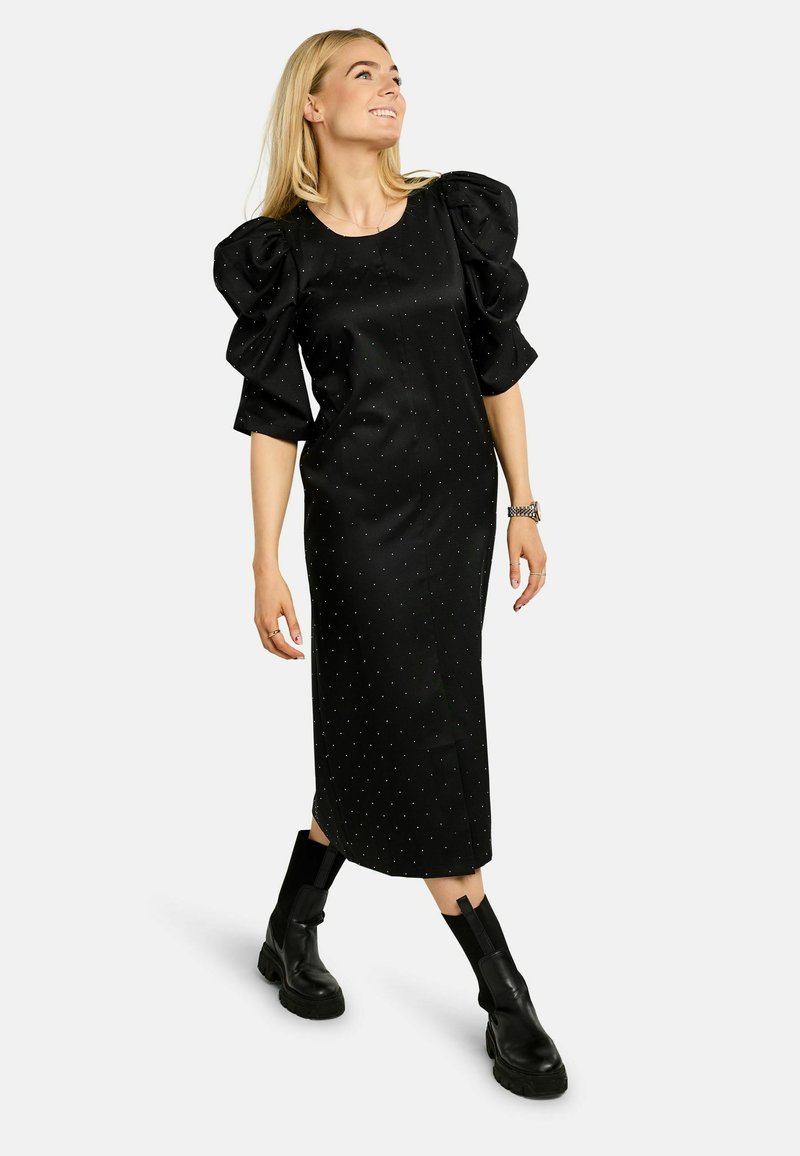 Rochie neagră cu mâneci bufante, cu model cu buline, până la genunchi. Purtați cu ghete negre până la gleznă. Material neted cu o siluetă ajustată.