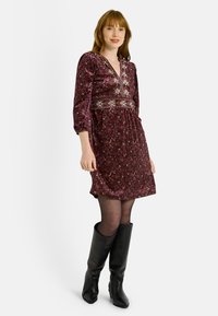 Vestido floral burdeos con escote en V, con bordados intrincados en la cintura, mangas largas y un corte relajado; combinado con botas negras hasta la rodilla.