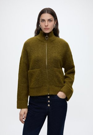 Femme portant un pull zippé vert olive avec de grandes poches et un jean taille haute bleu foncé avec quatre boutons devant, sur un fond uni.