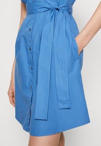 Blauwe korte jurk gemaakt van lichtgewicht materiaal, met een knoopsluiting aan de voorkant, een afgeronde zoom en een tailleceintuur die gestrikt kan worden. Voorzien van zijzakken.