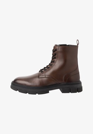 Bottine en cuir marron avec des lacets noirs, bout rond et semelle en caoutchouc épaisse. Présente des détails de couture et une languette à l'arrière.