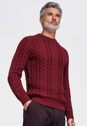 Homme aux cheveux gris et à la barbe portant un pull torsadé rouge foncé texturé et un pantalon sombre, les mains dans les poches.