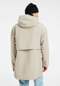 Giacca impermeabile beige con un design posteriore a strati, cappuccio regolabile e lati con cerniera. Abbinata a jeans azzurri chiari e un berretto nero.