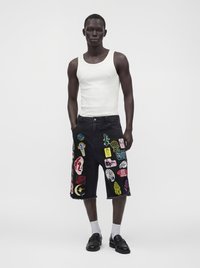 Man wearing white sleeveless top, black knee-length shorts with colorful patches, white socks, and black loafers standing against plain background.