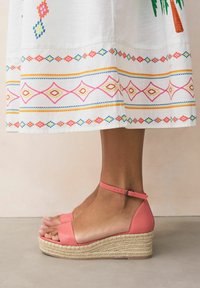 Korall sz�ínű wedge szandál texturált espadrille talppal. Sarokpánttal és széles lábujjpánttal rendelkezik, egy fehér hímzett szoknya mellett.