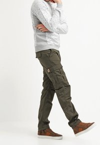 Oliwkowe spodnie typu cargo z licznymi kieszeniami, wykonane z wytrzymałej tkaniny, zestawione z jasnoszarym, wzorzystym swetrem oraz brązowymi tenisówkami.