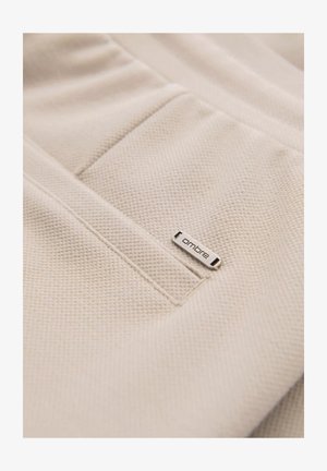 Beige textiel met een gestructureerd oppervlak; heeft een klein zilveren merketiket met de tekst "ombre" en een genaaide zakdetail.