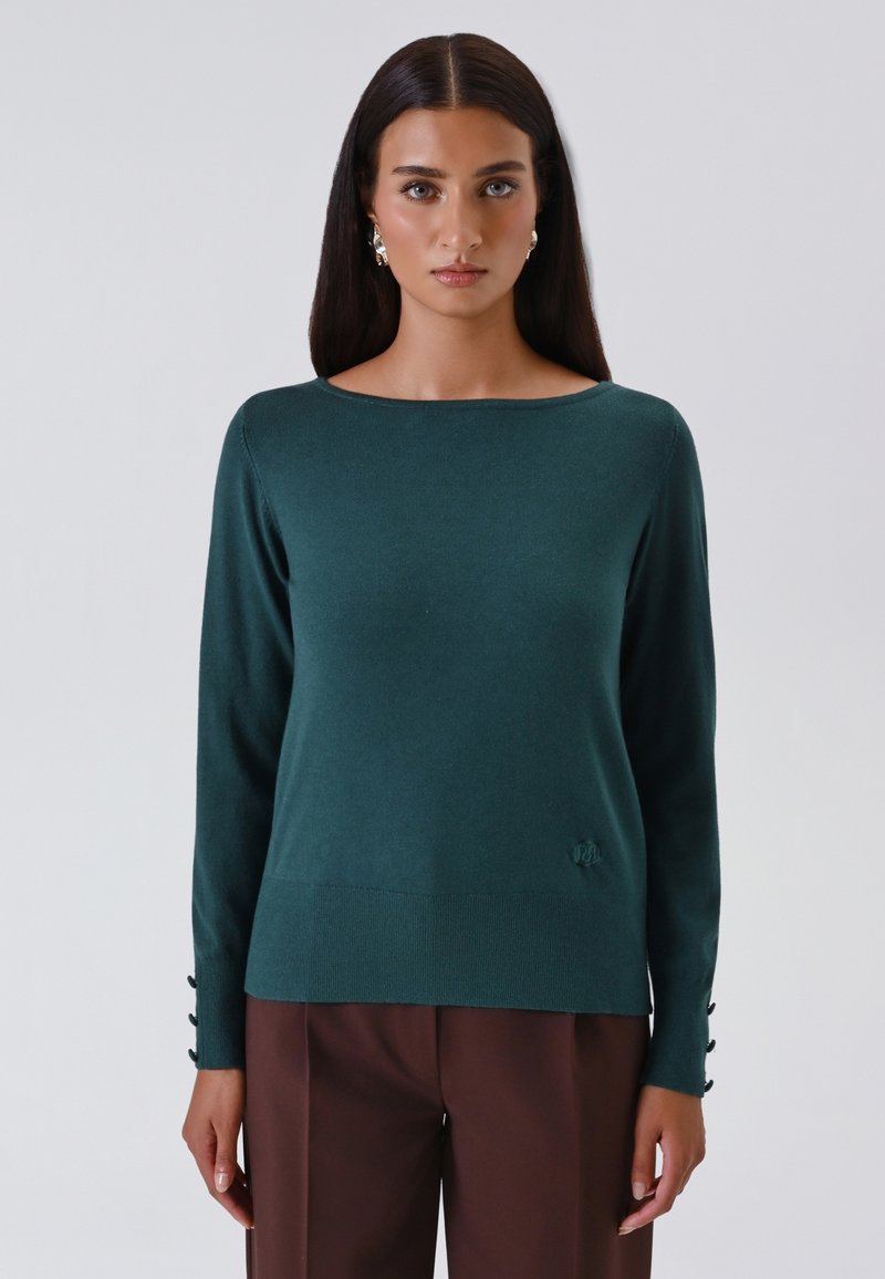 Vrouw met steil zwart haar, gekleed in een teal lange mouwen trui met knoopmanchetten en een bruine broek, staand tegen een effen lichte achtergrond.