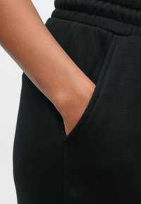 Juodi sportiniai kelnių su elastinga juosmens juosta, šonine kišene ir lygia tekstūra, ranka atsirėmusi į kišenę.