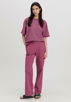 Pink cotton oversized t-shirt and matching loose pants. Smooth texture, relaxed fit, and subtle logo detail on the shirt. Black sandals.