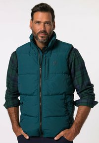 Teal puffer vest met een rits aan de voorkant, hoge kraag en zijzakken. Gedragen over een groen en marineblauw geruit shirt met een logo detail.