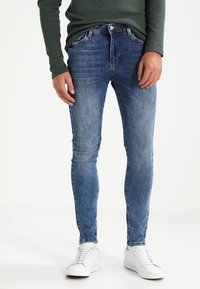 Slim-fit siniset farkut, joissa on haalistunut pesu, viisi taskua ja napitus, yhdistettynä valkoisiin lenkkareihin ja vihreään puseroon.