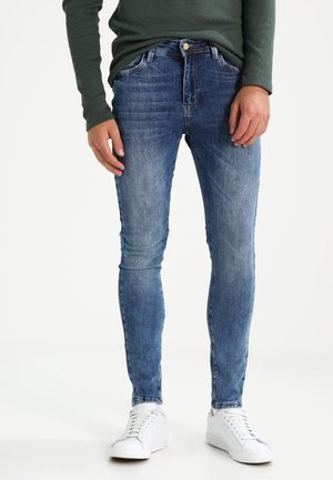 Slim-fit blue denim jeans with a faded wash, five-pocket design, and a button closure, paired with white sneakers and a green top.