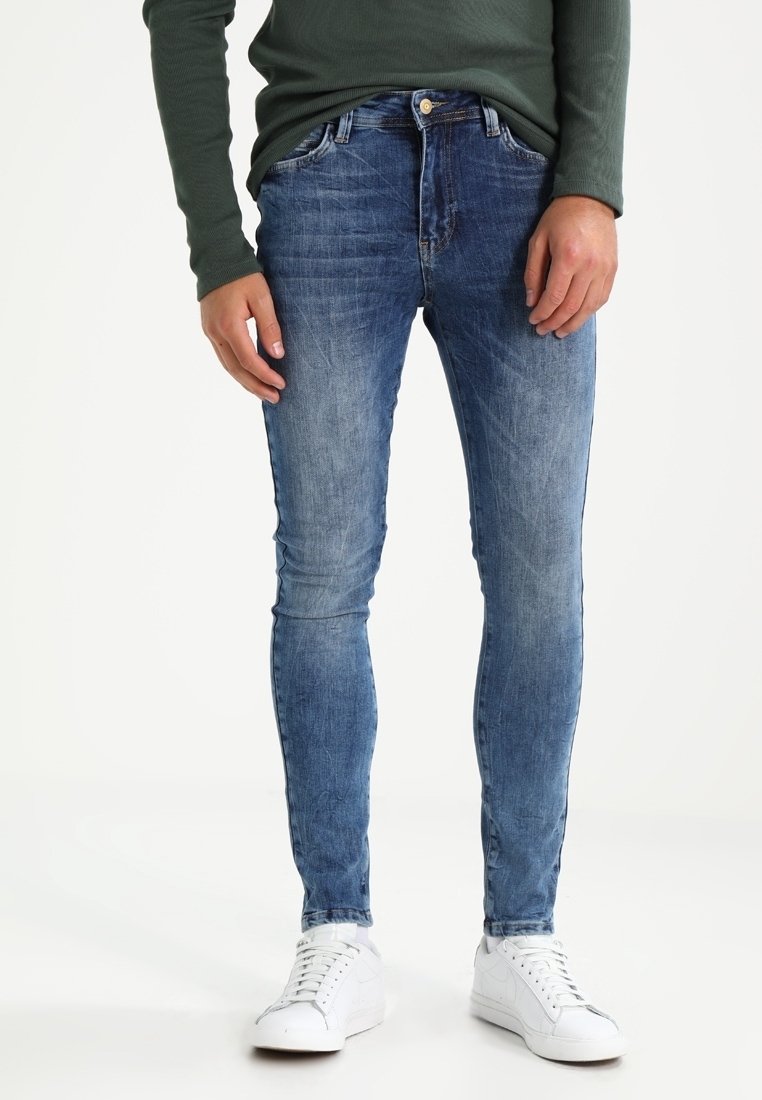 Slim-fit siniset farkut, joissa on haalistunut pesu, viisi taskua ja napitus, yhdistettynä valkoisiin lenkkareihin ja vihreään puseroon.