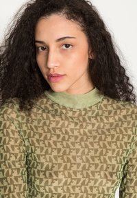 Blusa de mangas compridas em tecido transparente e texturizado, com uma base de cor verde oliva e um padrão geométrico. Decote alto e design ajustado.