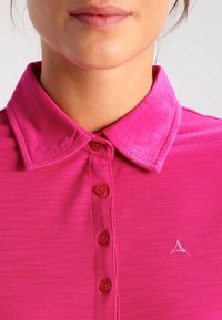 Polo rose en tissu texturé, avec un col et cinq boutons. Un petit logo est brodé sur le côté gauche.