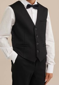 Svart väst med en strukturerad yta, treknappsstängning, två framfickor och spetsig fåll, i kombination med en vit skjorta och fluga.