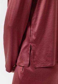 Bordeaux langemouwtop met een relaxte fit, zachte textuur, zijsplitten en rechte zoom. De stof lijkt lichtgewicht en glad.