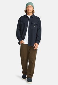 Marineblauw flanellen shirt met twee borstzakken, gecombineerd met bruine cargobroek en marineblauwe sneakers. Een groene gebreide beanie maakt de outfit compleet.