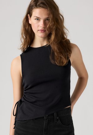 Jeune femme aux cheveux bruns ondulés, portant un top noir sans manches et un jean noir, debout avec une main sur la hanche devant un fond uni.