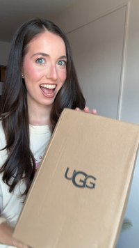 Brown cardboard box with UGG logo, held by a person with long hair. The box has a rectangular shape and smooth texture.