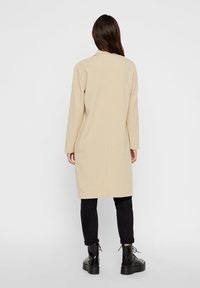 Beige dlouhý kabát s rovnou siluetou a dlouhými rukávy, vyrobený z hladkého materiálu, v kombinaci s černými zúženými kalhotami a masivními černými botami.